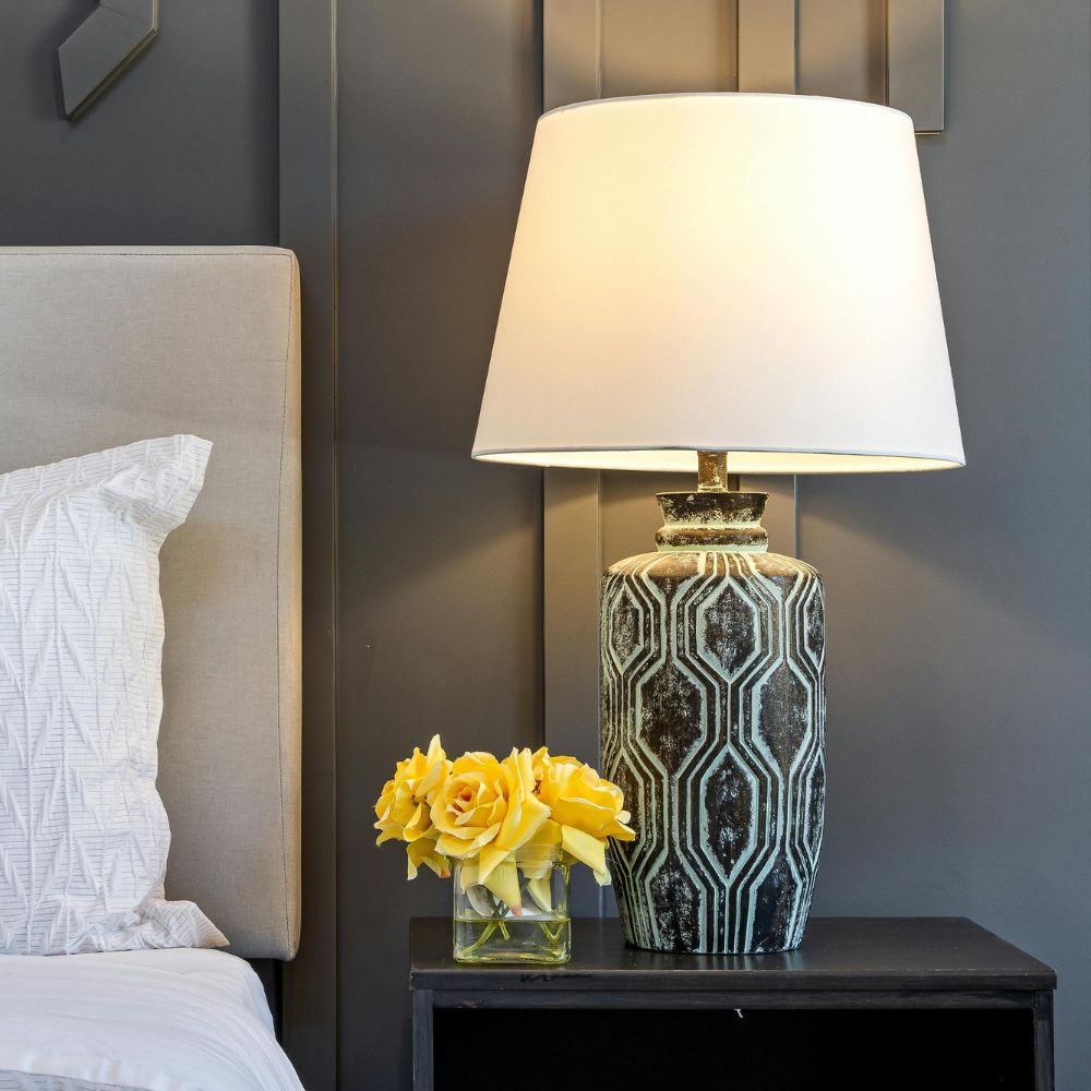 Decorative lamp on a nightstand with yellow flowers next to a bed in a bedroom setting.